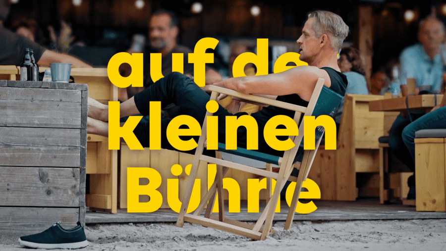 Filmszene mit Publikum vor kleiner Festivalbühne und integrierter gelber Typografie.